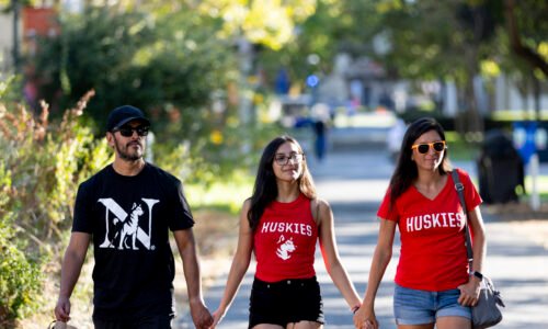 Three people walk along a sunlit, tree-lined campus path, wearing Northeastern branded t-shirts. Academic buildings and other pedestrians are blurred, but visible in the background.