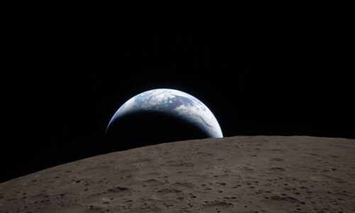 art002e009288 (April 6, 2026) – Earthset captured through the Orion spacecraft window at 6:41 p.m. EDT, April 6, 2026, during the Artemis II crew’s flyby of the Moon. A muted blue Earth with bright white clouds sets behind the cratered lunar surface. The dark portion of Earth is experiencing nighttime. On Earth’s day side, swirling clouds are visible over the Australia and Oceania region. In the foreground, Ohm crater has terraced edges and a flat floor interrupted by central peaks. Central peaks form in complex craters when the lunar surface, liquefied on impact, splashes upwards during the crater’s formation.