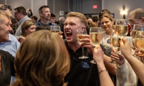A group of people raise glasses of white wine in a celebratory toast, with one man in the center smiling and cheering enthusiastically in a crowded indoor setting.
