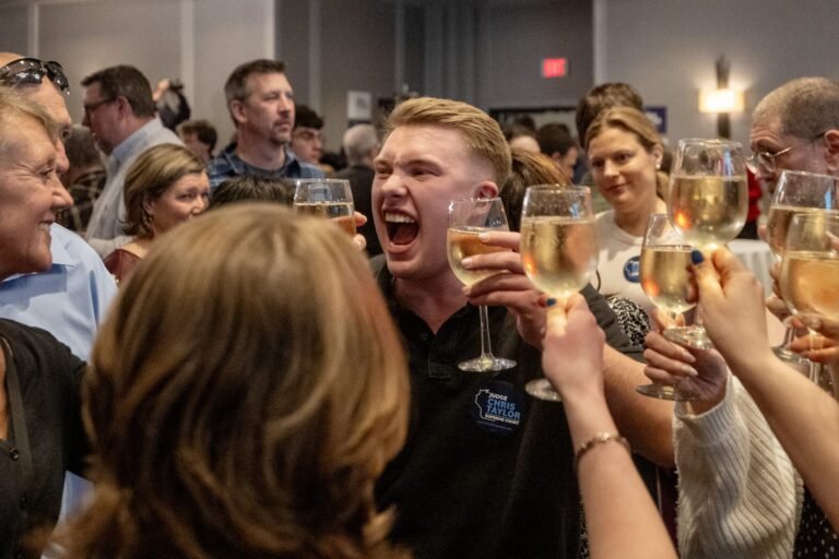 A group of people raise glasses of white wine in a celebratory toast, with one man in the center smiling and cheering enthusiastically in a crowded indoor setting.