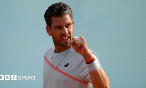 Cameron Norrie is wearing a white t-shirt with a red sash and holding up his fist