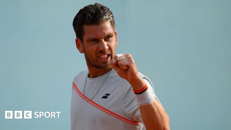Cameron Norrie is wearing a white t-shirt with a red sash and holding up his fist