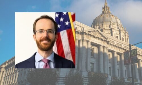 A man in a suit and tie stands in front of an American flag; in the background is a large government building with a dome and ornate architecture.