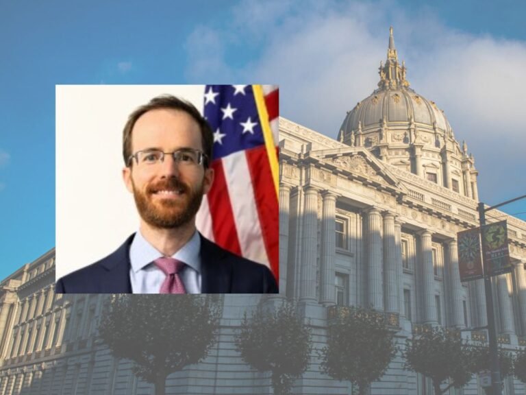 A man in a suit and tie stands in front of an American flag; in the background is a large government building with a dome and ornate architecture.