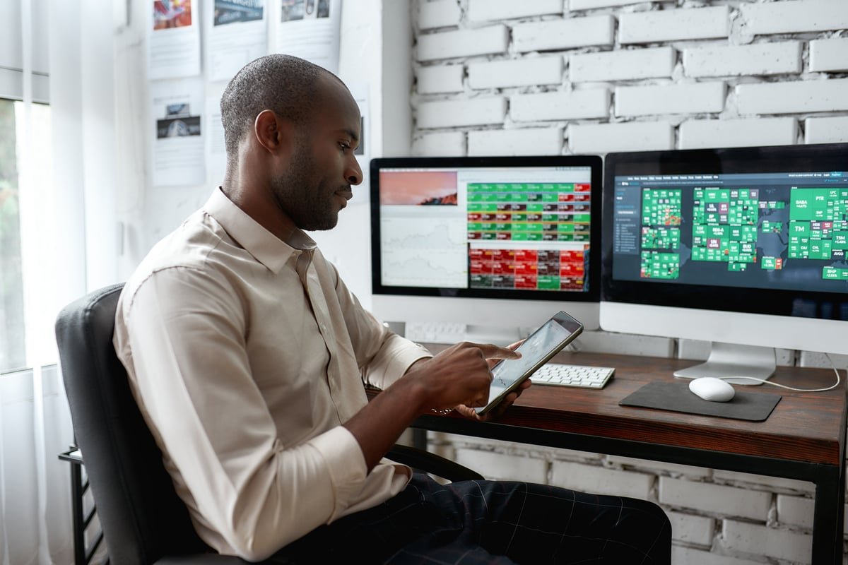 An investor looking at their smartphone with computer screens in the background, showing stock prices.