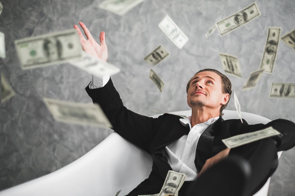 Person in a suit sitting in a bathtub amid flying currency notes.