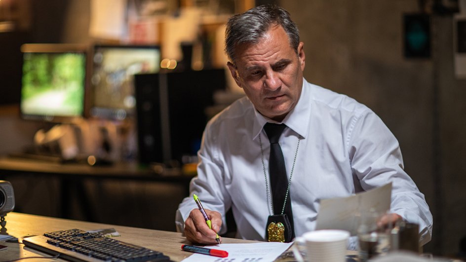 Gray-haired police officer in white shirt working late at night in the office, sitting at desk, looking at photograph and writing