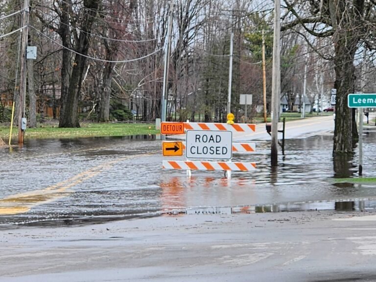 Flooded street with Road Closed and detour signs blocking vehicle access; water covers the road and spills onto the grass.