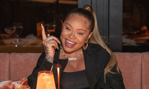 Latto, with long hair in a ponytail, smiles while sitting at a table with a drink, wearing a black outfit and jewelry.