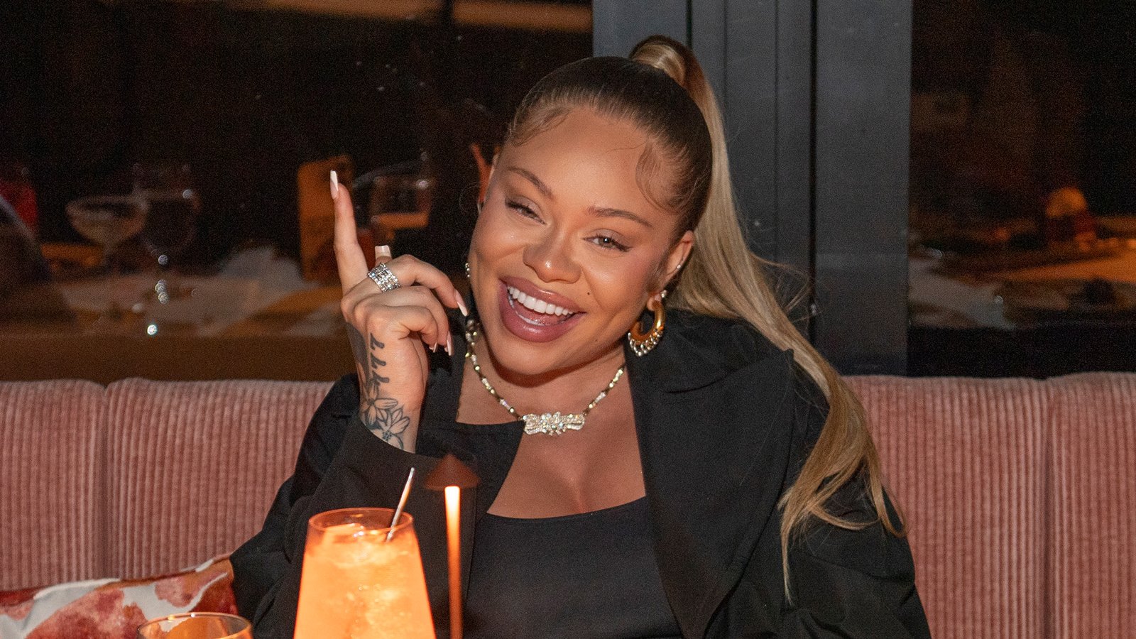 Latto, with long hair in a ponytail, smiles while sitting at a table with a drink, wearing a black outfit and jewelry.