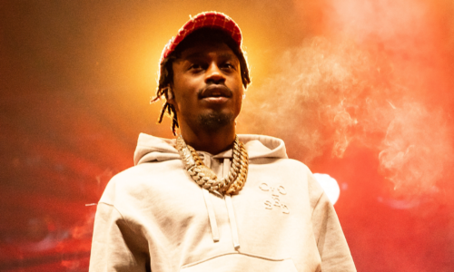 Lil Tjay in a hoodie and cap is performing on stage, with a smoky, colorful background.