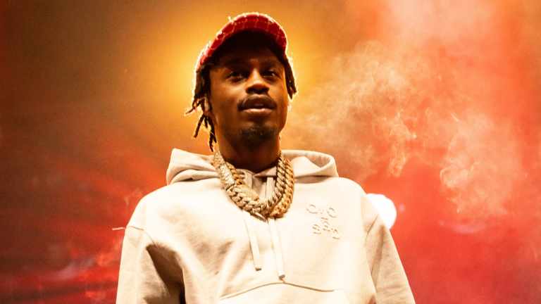 Lil Tjay in a hoodie and cap is performing on stage, with a smoky, colorful background.