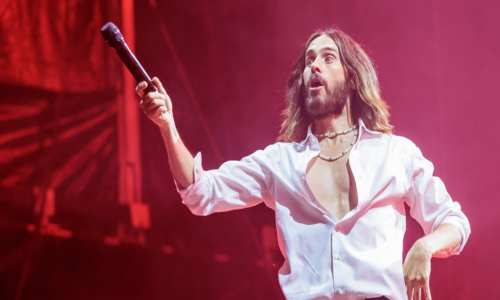 A man with long hair and a beard, wearing an open white shirt and black pants, holds a microphone extended in one hand on a red-lit stage.