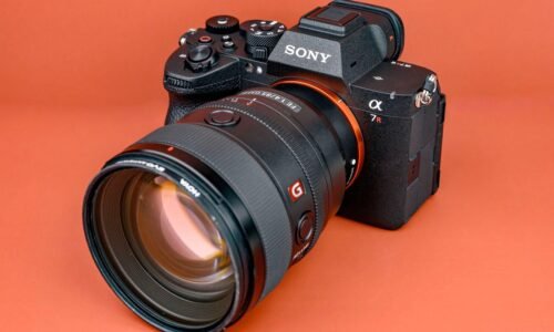 A Sony Alpha 7R mirrorless camera with a large zoom lens attached, placed on an orange surface with visible control dials and logos.