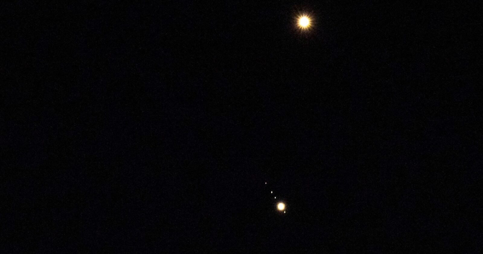 Venus is pictured shining brightly above Jupiter in a black sky. A number of small star-like objects can be seen in a line surrounding Jupiter, representing its four largest Galilean Moons.