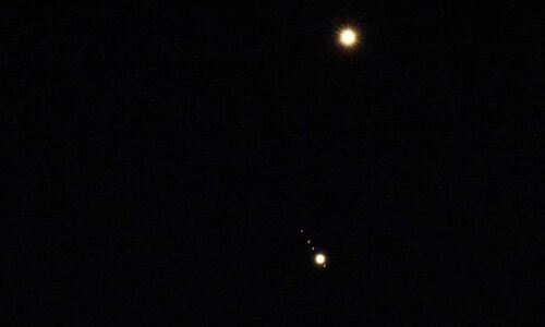 Venus is pictured shining brightly above Jupiter in a black sky. A number of small star-like objects can be seen in a line surrounding Jupiter, representing its four largest Galilean Moons.