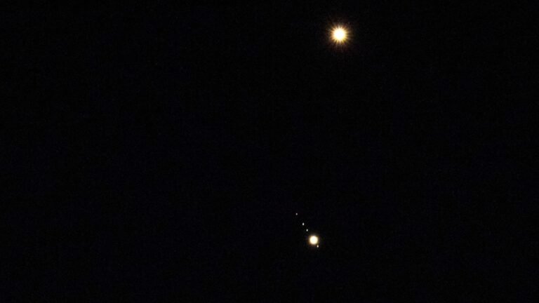 Venus is pictured shining brightly above Jupiter in a black sky. A number of small star-like objects can be seen in a line surrounding Jupiter, representing its four largest Galilean Moons.
