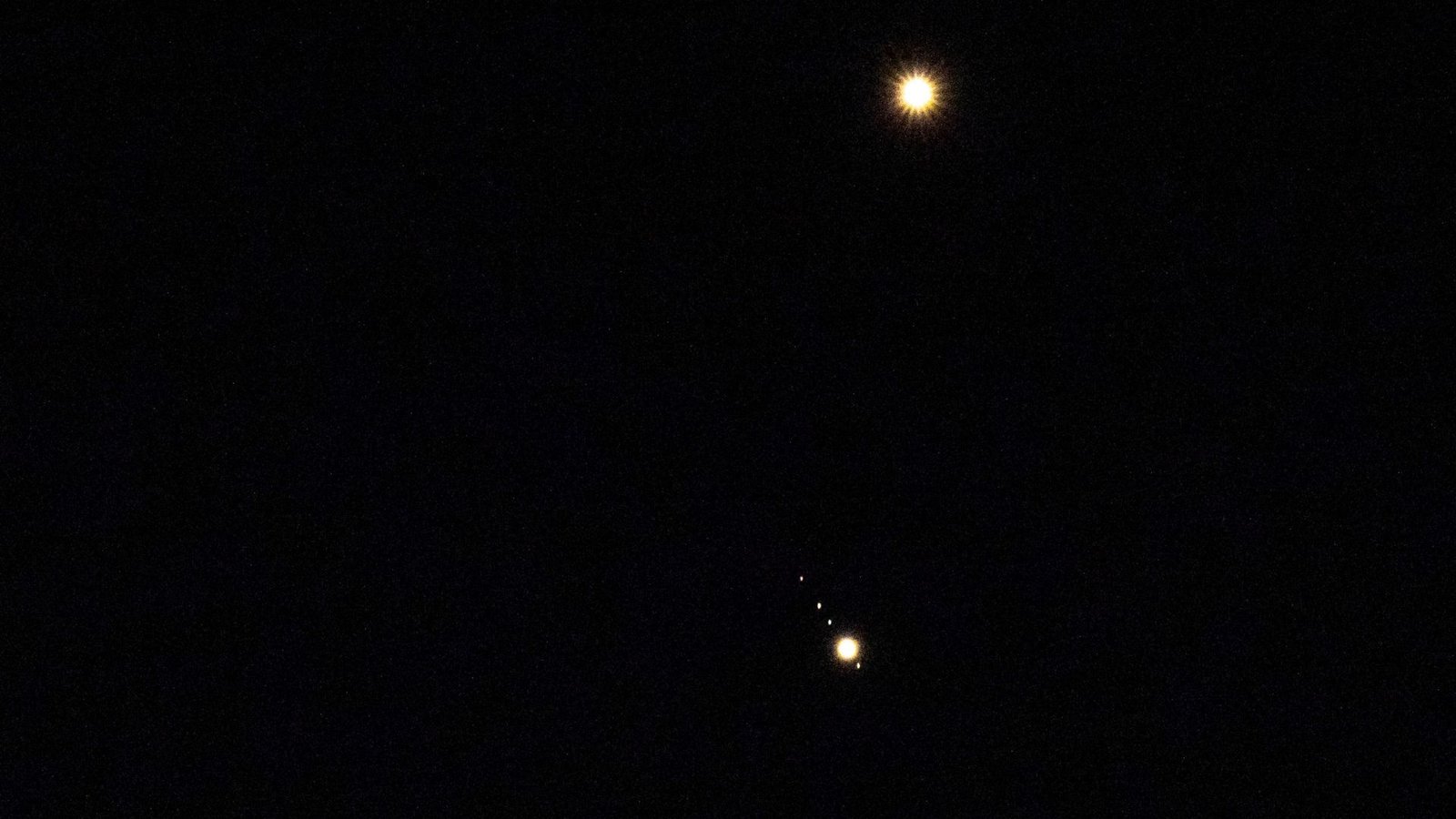 Venus is pictured shining brightly above Jupiter in a black sky. A number of small star-like objects can be seen in a line surrounding Jupiter, representing its four largest Galilean Moons.