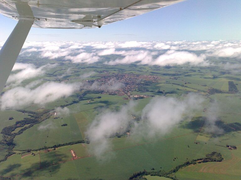 Vista aérea de Ubiratã, no Paraná, entre lavouras verdes, com nuvens baixas e a asa de um avião em primeiro plano