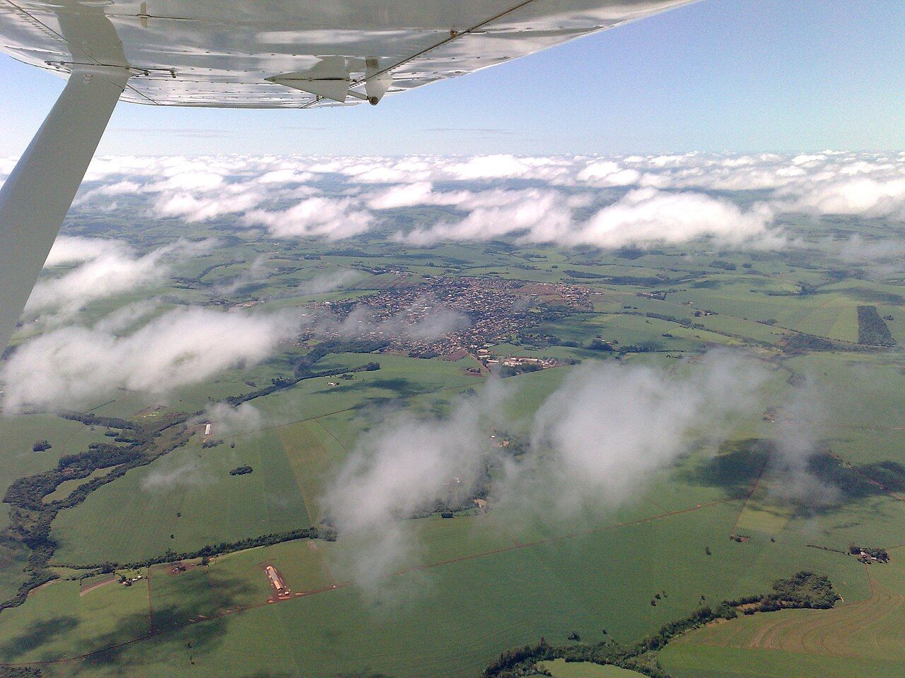 Vista aérea de Ubiratã, no Paraná, entre lavouras verdes, com nuvens baixas e a asa de um avião em primeiro plano